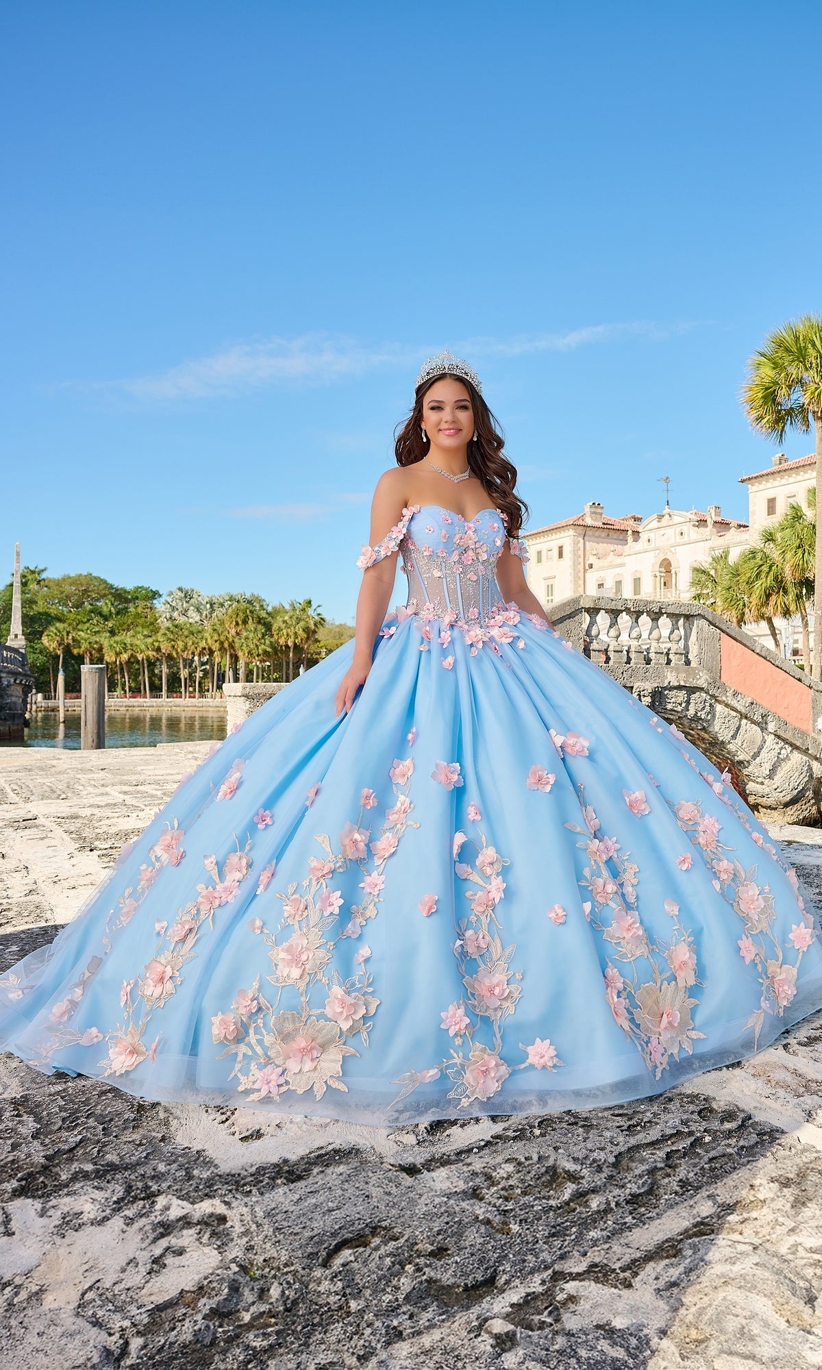 A young woman in the Quinceanera Dress 54224 by Amarra, featuring off-the-shoulder pink floral details and a tiara, stands outdoors near stone steps and palm trees under a clear sky.